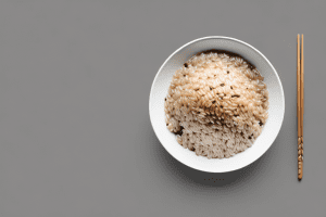 A bowl of steaming brown rice with a spoon and a few grains of rice scattered around it